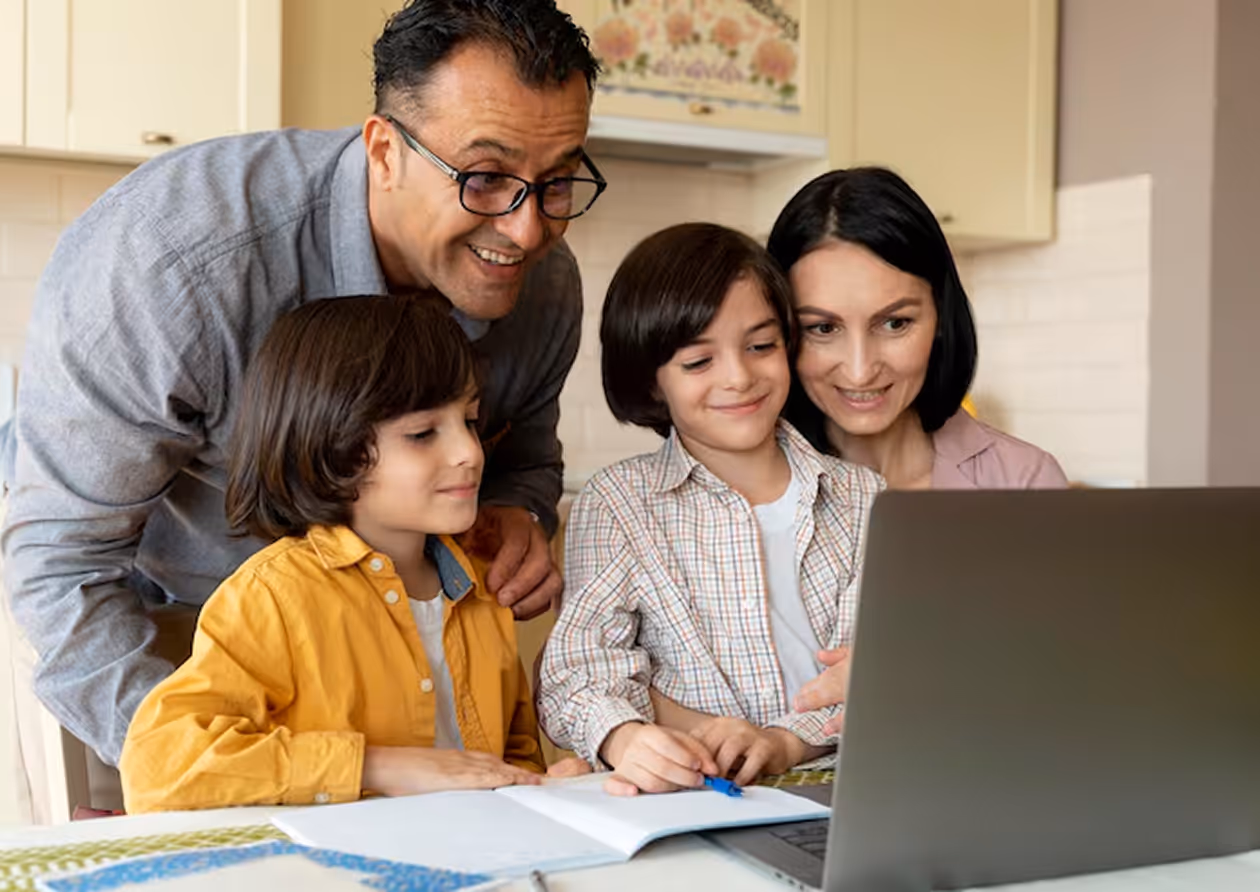 Smiling family of four gathered around a laptop at a table with notebooks, engaged in learning or browsing.