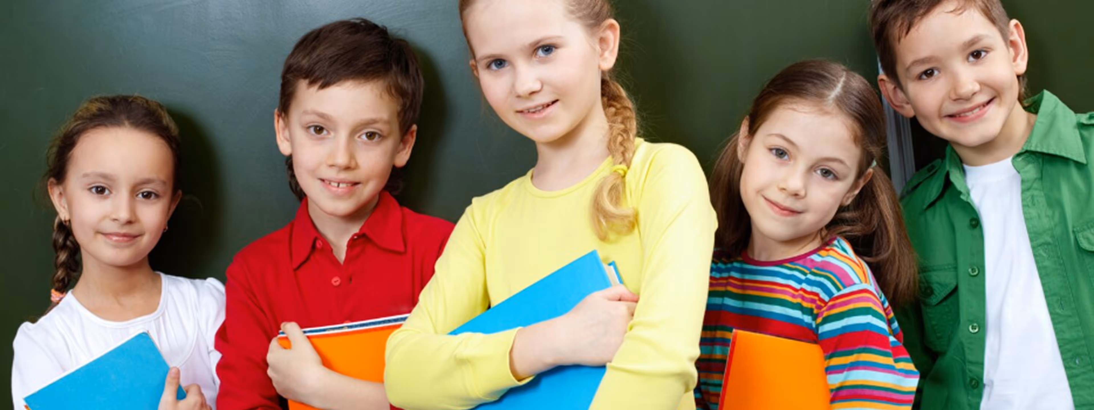 Five school children standing in front of a chalkboard, holding colorful notebooks and smiling at the camera.