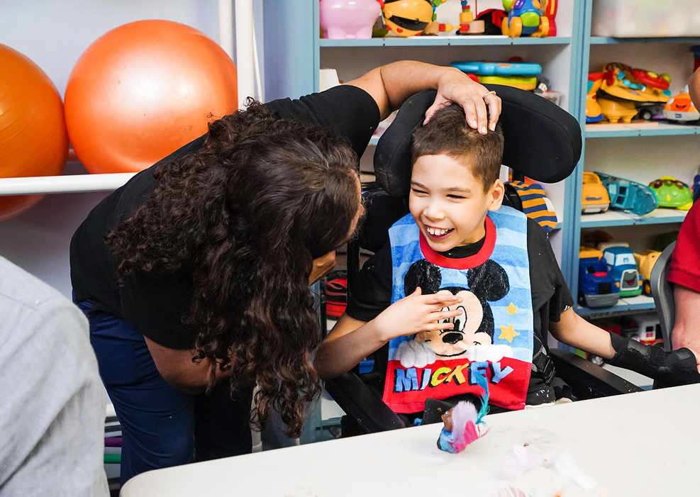 A smiling boy in a wheelchair wearing a Mickey Mouse bib, with a woman bending down and gently touching his head in a playroom with toys and orange balls in the background.