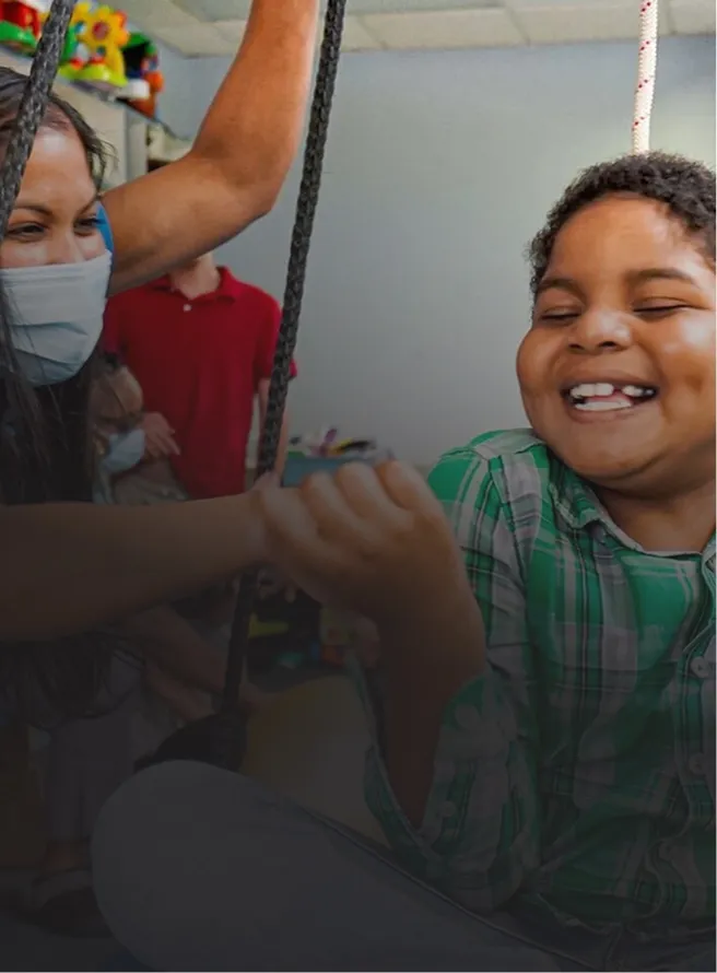 Smiling child in a green plaid shirt holding a rope swing indoors, assisted by a masked adult.