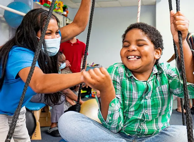 Smiling child in a green plaid shirt sitting on a swing being gently pushed by a masked adult in a blue shirt inside a playroom.