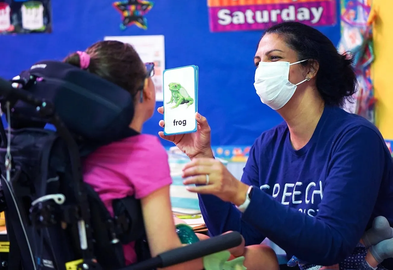 Speech therapist wearing a mask holding a flash card with a frog image to a child in a wheelchair.
