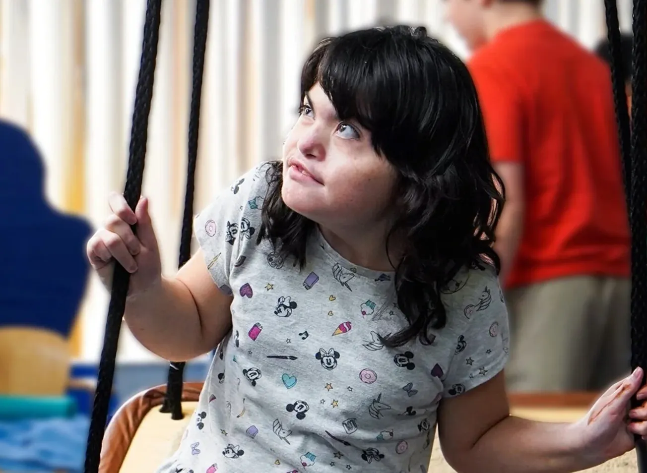 Girl with dark hair wearing a patterned t-shirt sitting on a swing, looking upward thoughtfully.