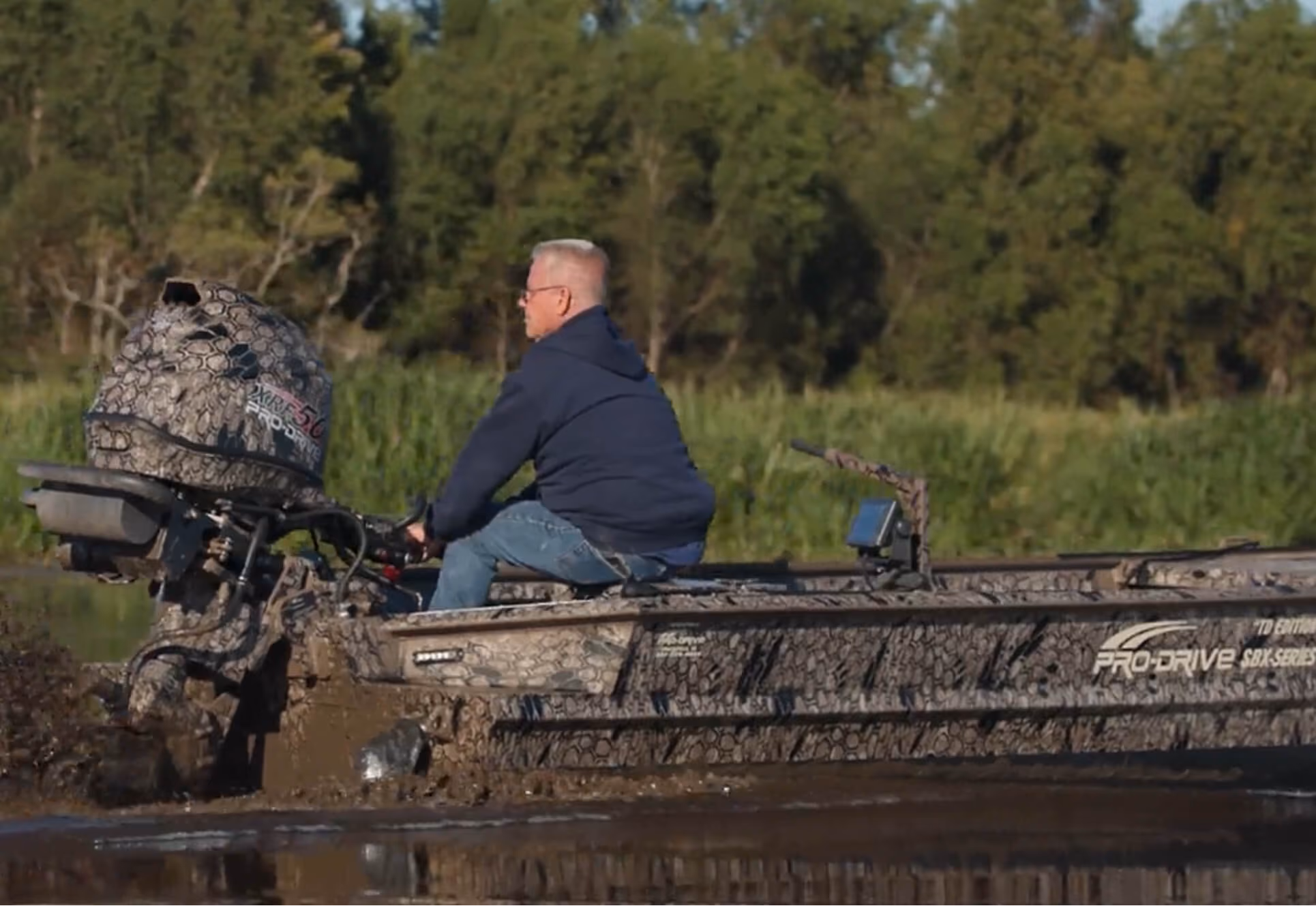 Man wearing a navy hoodie and jeans driving a camouflage-patterned Pro-Drive boat through muddy water near green trees.