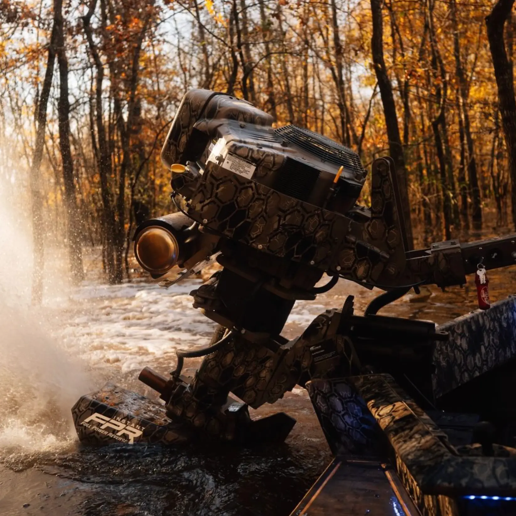 Outboard motor with camouflage pattern mounted on a boat splashing water in a wooded area with autumn foliage.