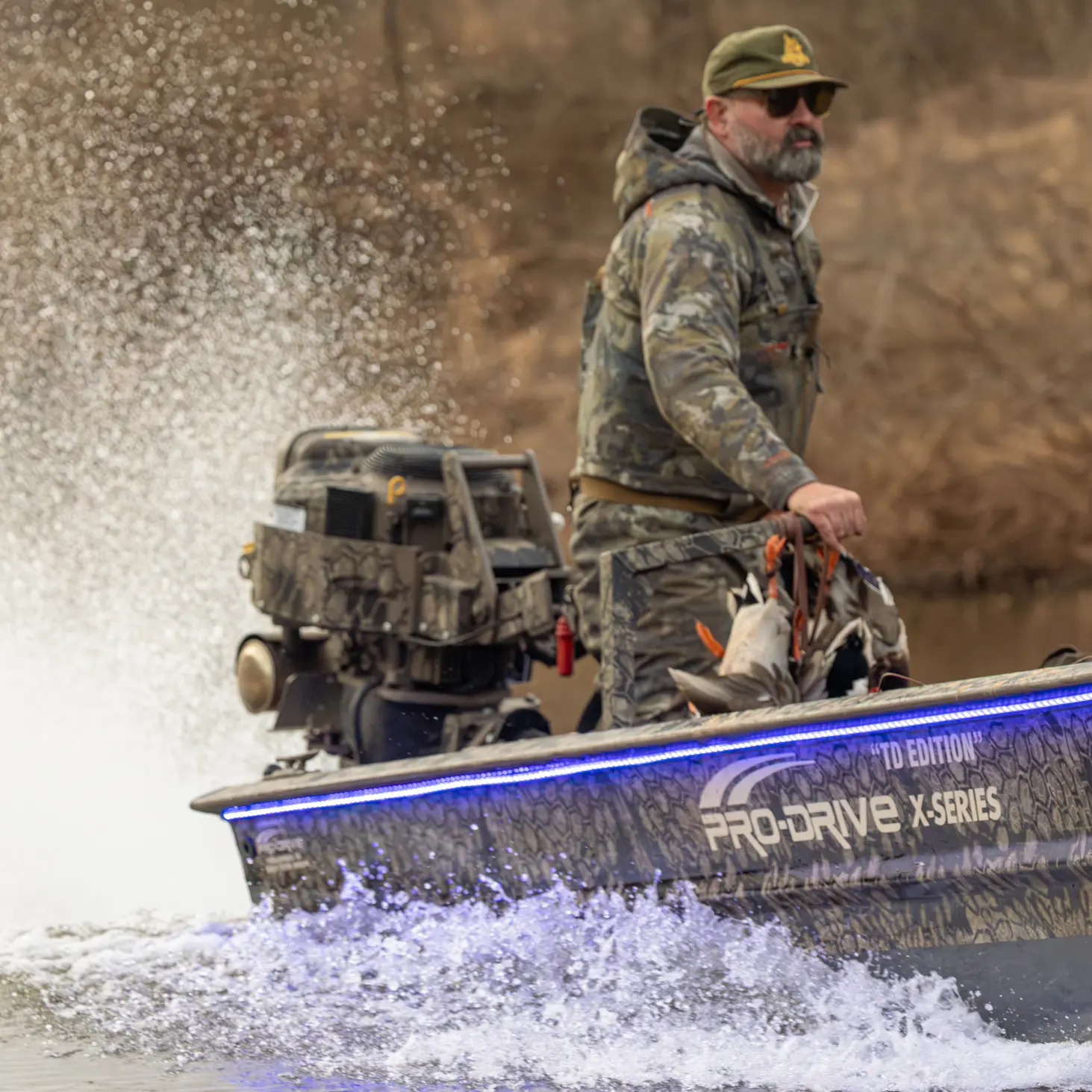 Man in camouflage clothing steering a camouflage Pro-Drive X-Series boat with water splashing behind.