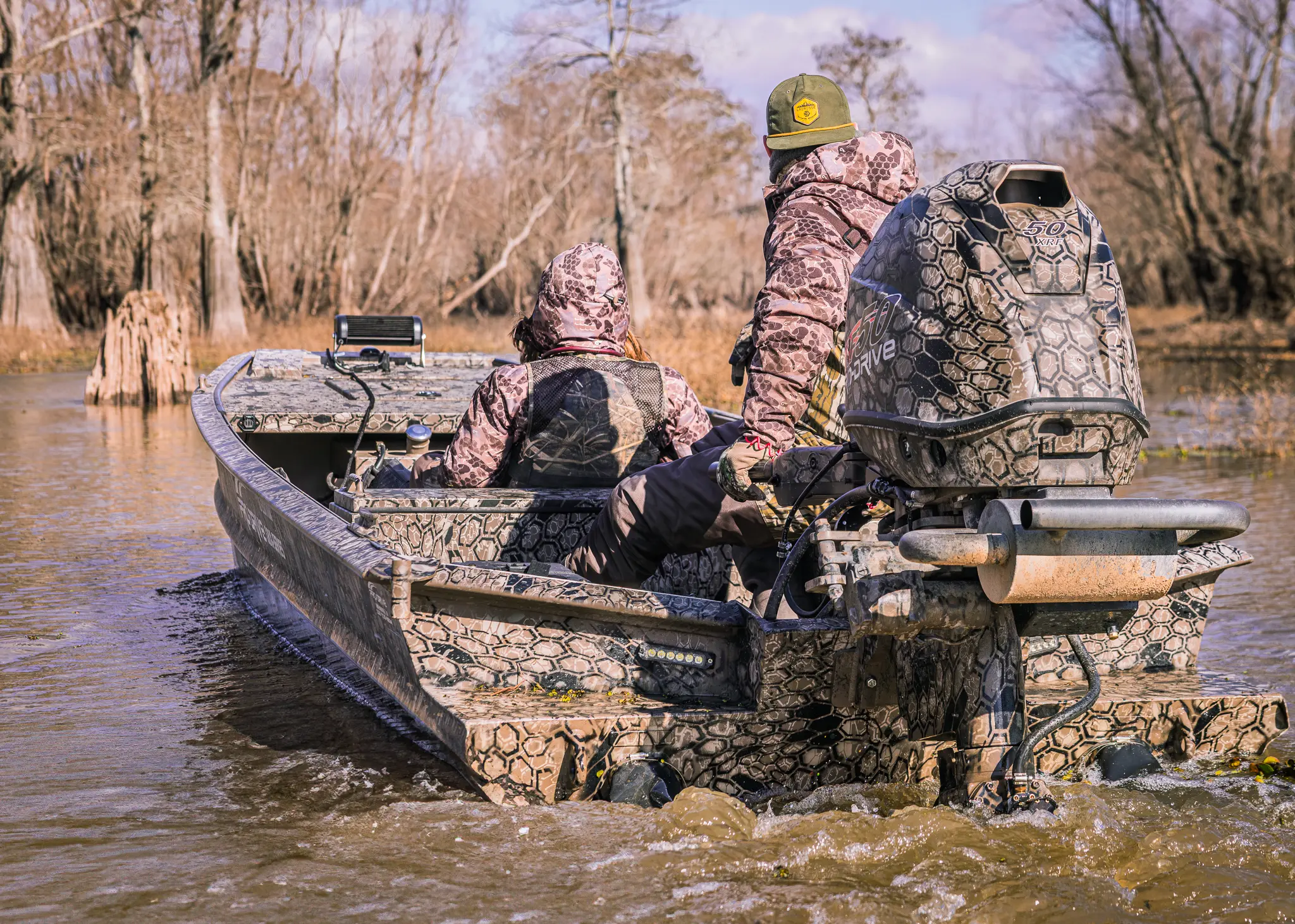 Two people in camouflage jackets on a camo-patterned boat with an outboard motor moving through shallow water surrounded by leafless trees.