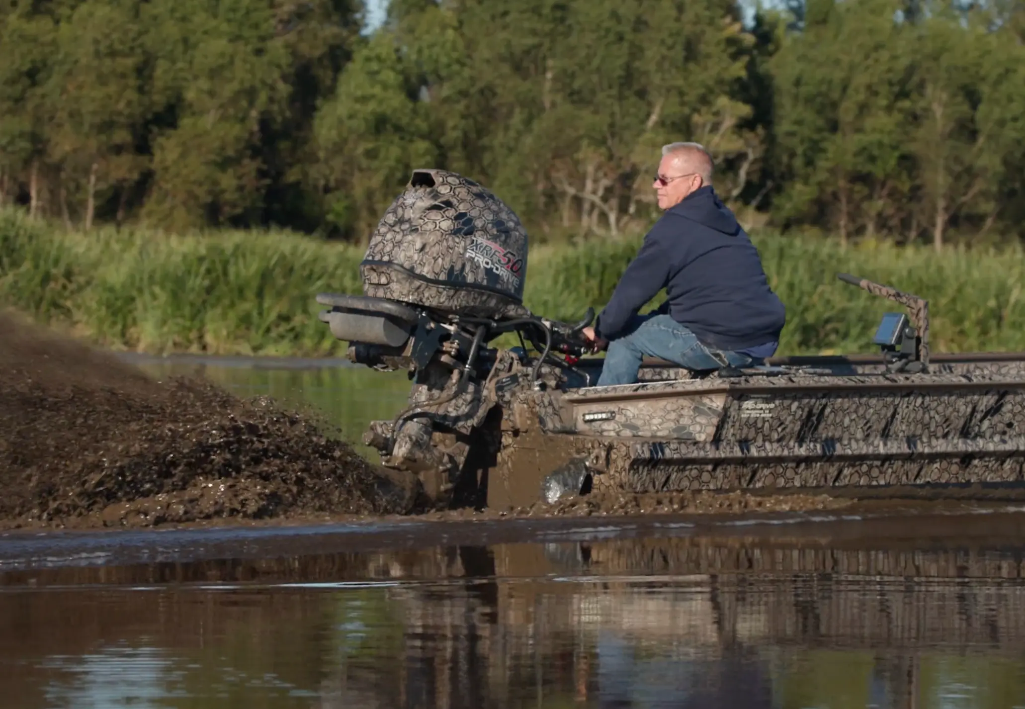 Man wearing sunglasses driving a camouflaged boat through shallow water splashing mud.