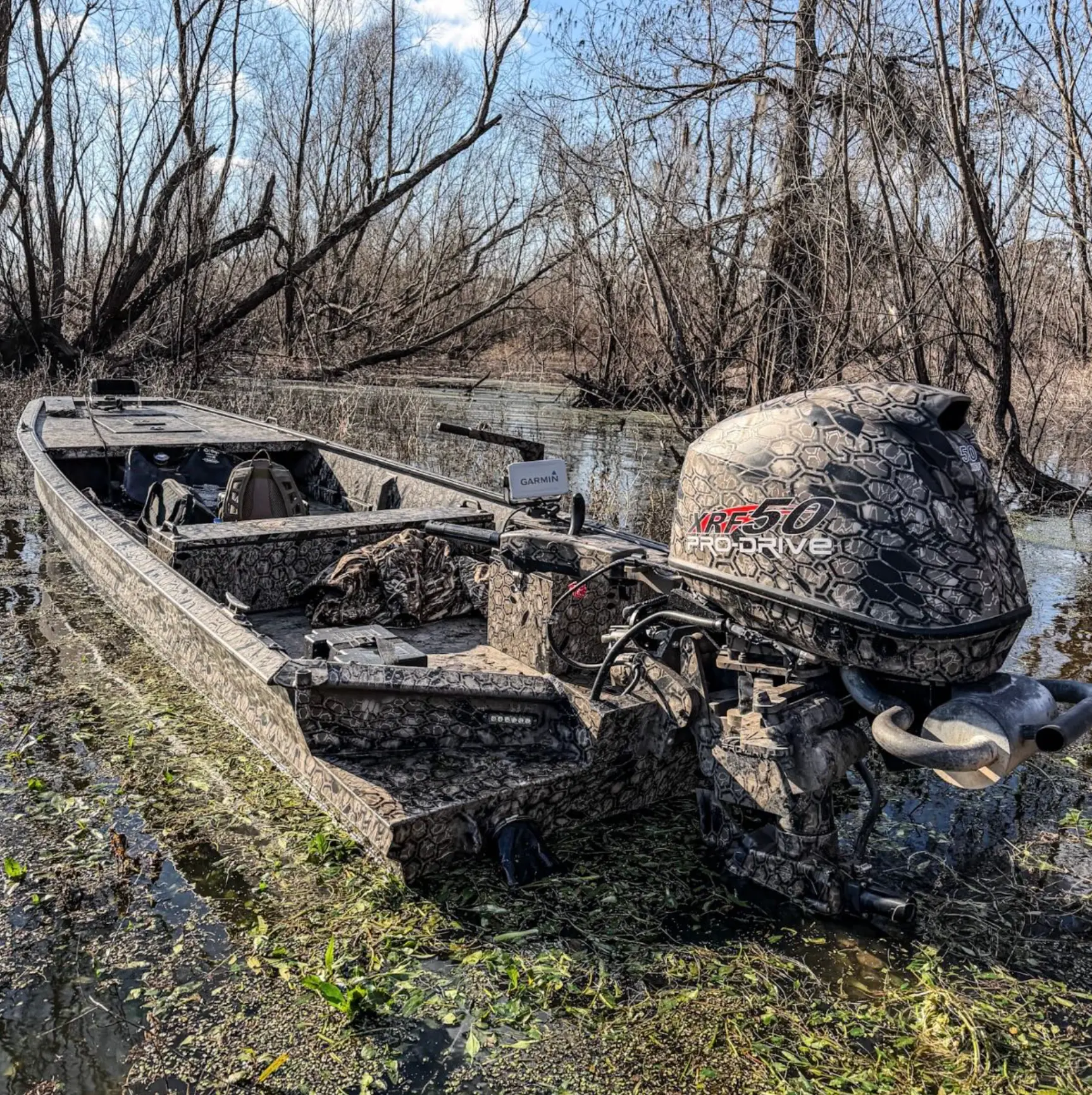 Camouflage-patterned XRF 50 Pro Drive boat equipped with a Garmin device in a shallow, overgrown water area with leafless trees.