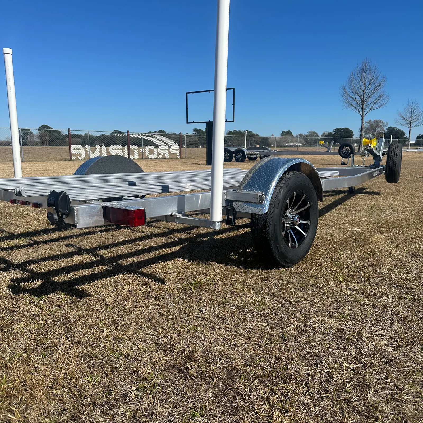 Metal boat trailer with black wheels parked on dry grass under a clear blue sky.