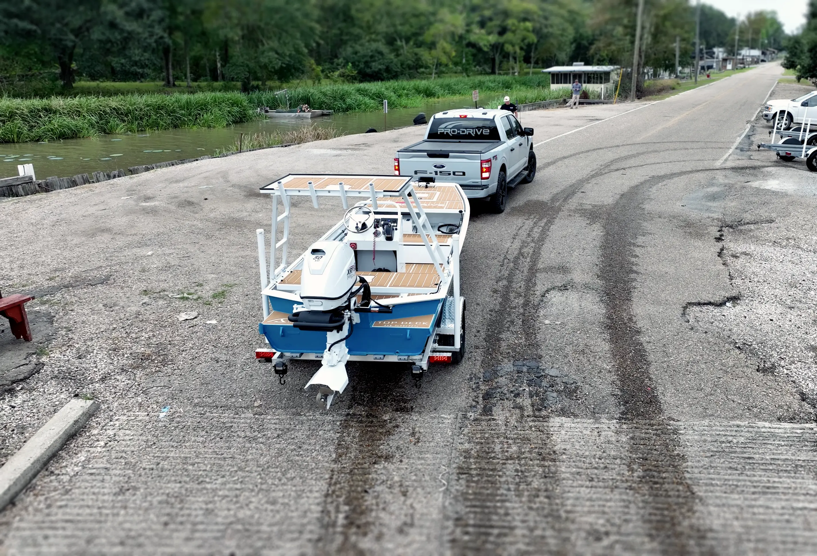 White Ford F-150 truck towing a blue and white motorboat on a trailer on a gravel boat ramp near a river.