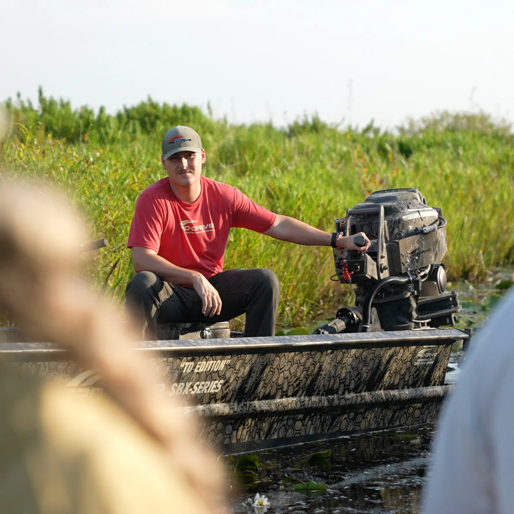Man wearing a red shirt and cap sitting in a camouflaged boat, holding the motor handle, surrounded by green vegetation.