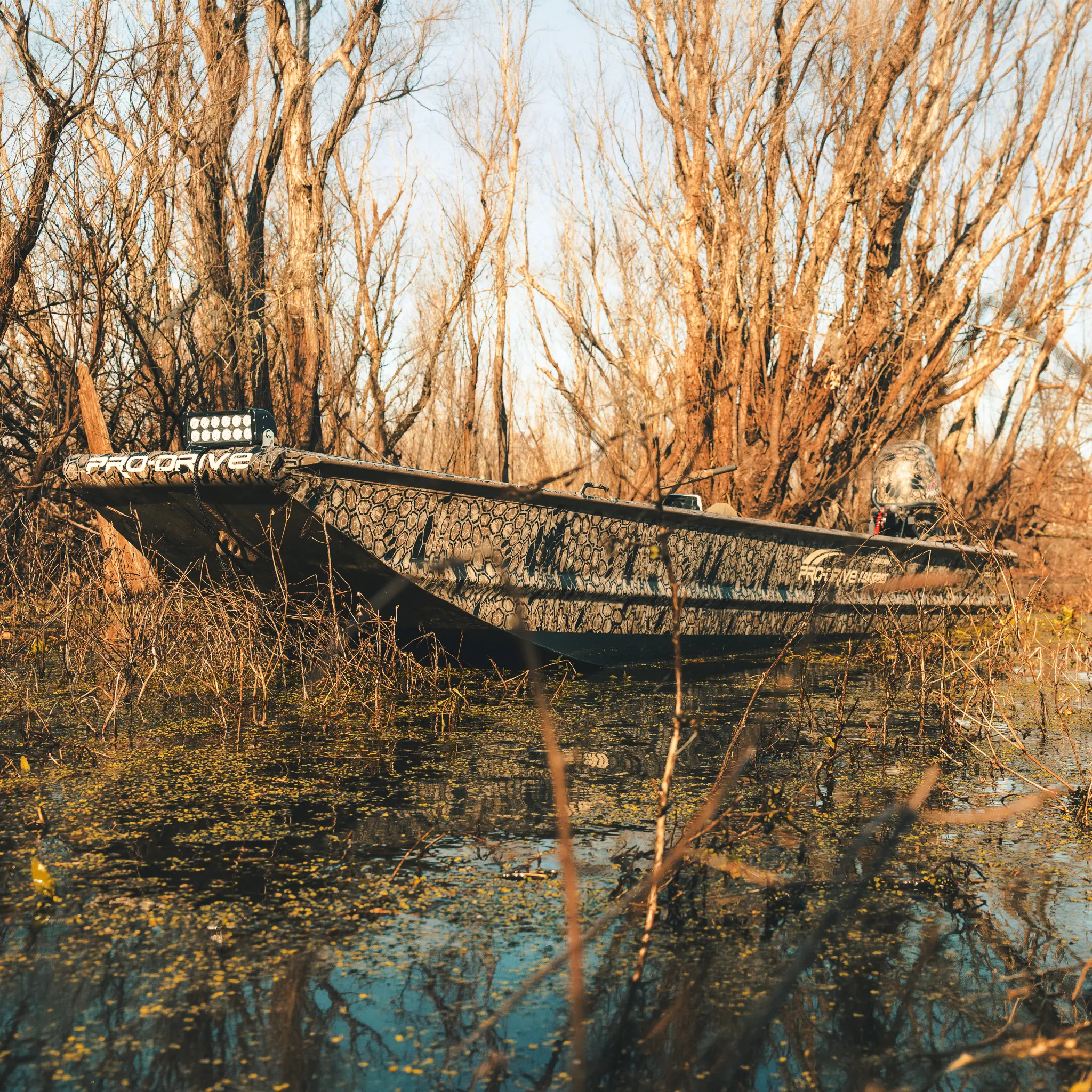Camouflage-patterned boat in shallow water surrounded by leafless trees and vegetation.