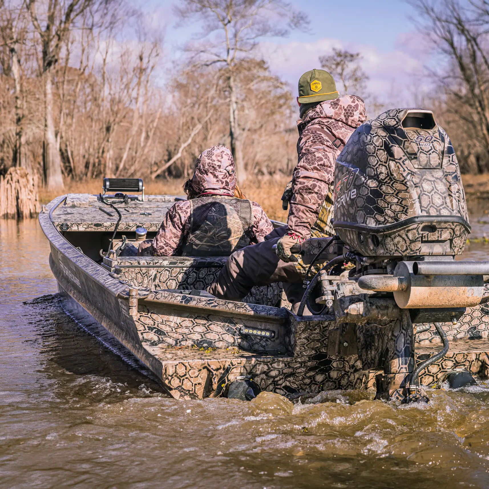 Two people wearing camo jackets riding a camouflaged motorboat through a shallow, tree-lined waterway.