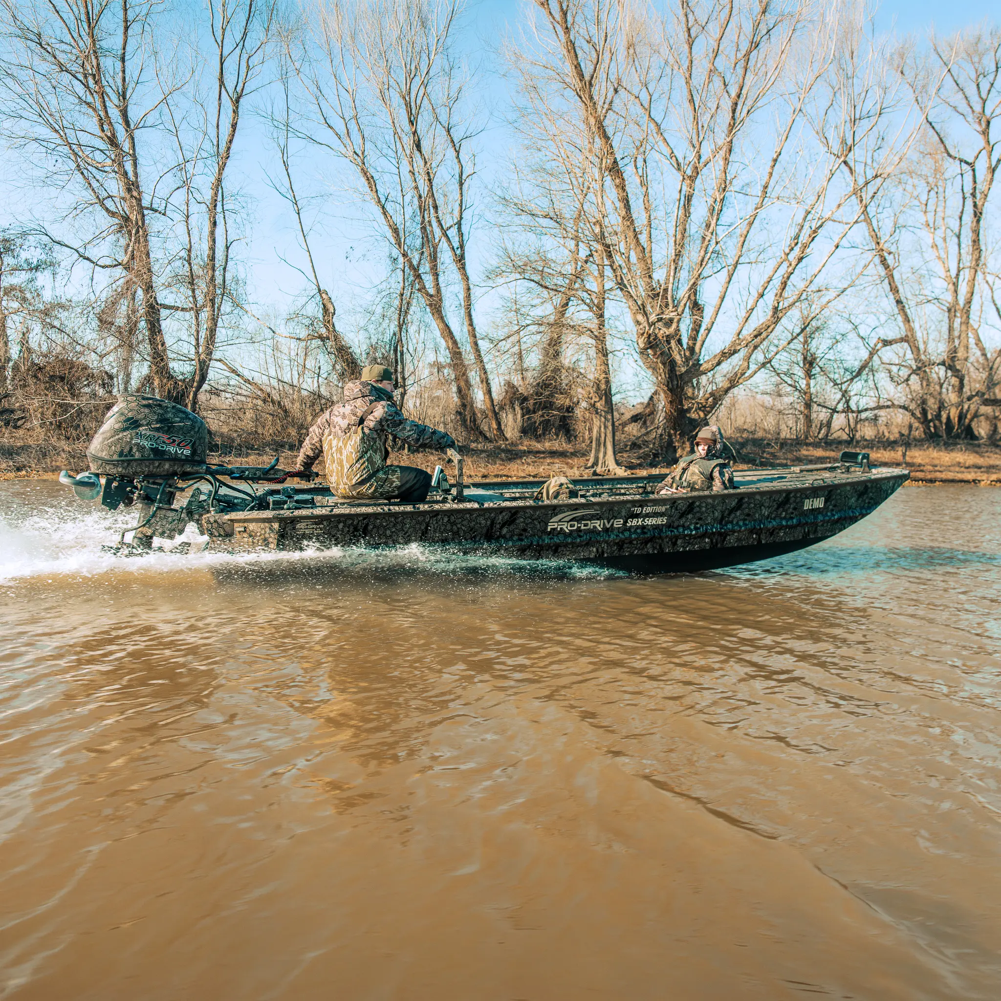 Two people in camouflage gear riding a camo-patterned motorboat on a river near leafless trees.