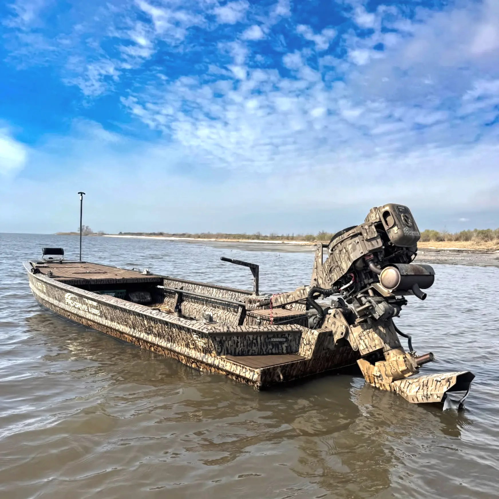 Camouflage-patterned flat-bottom boat with a motor floating on calm water near a shoreline under a partly cloudy blue sky.