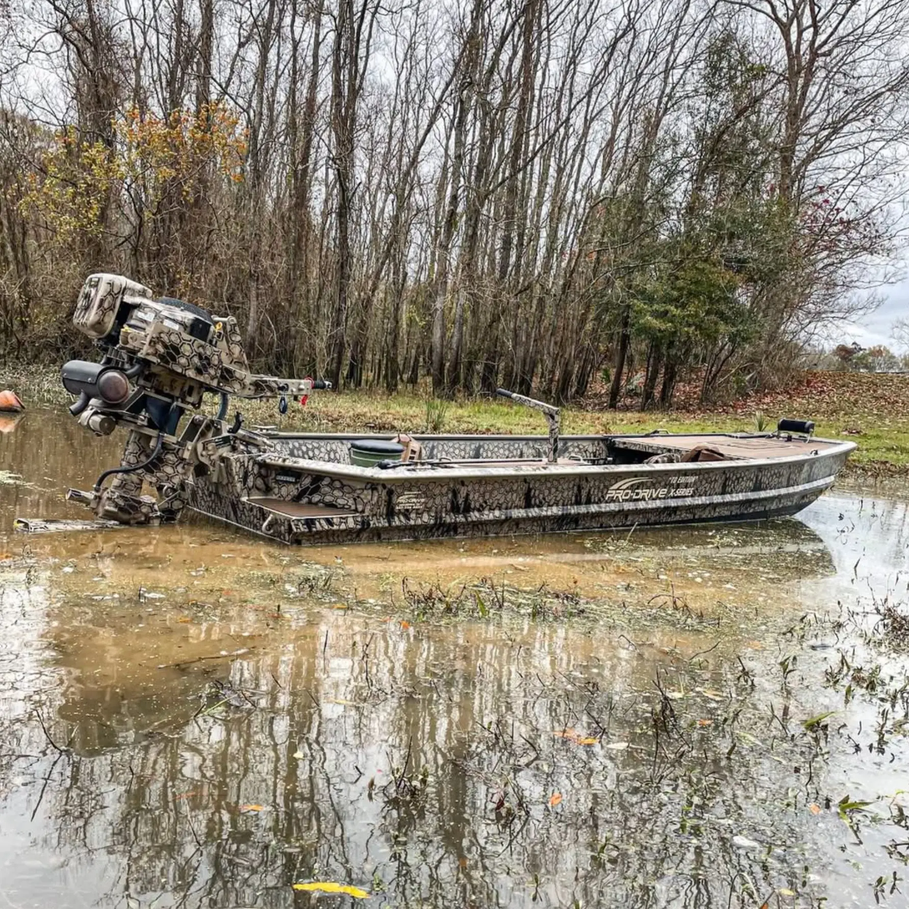 Camouflage Pro-Drive boat with motor in shallow water near a wooded area.