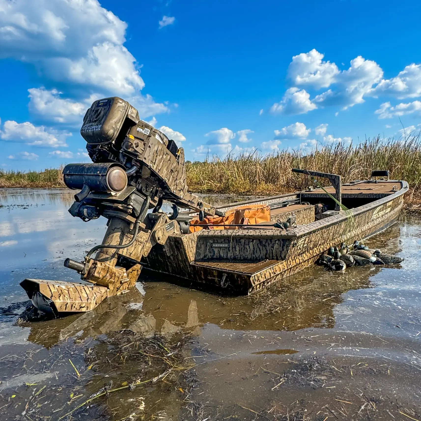 Camouflaged motorboat with outboard engine in shallow water near tall grass under a blue sky with clouds.