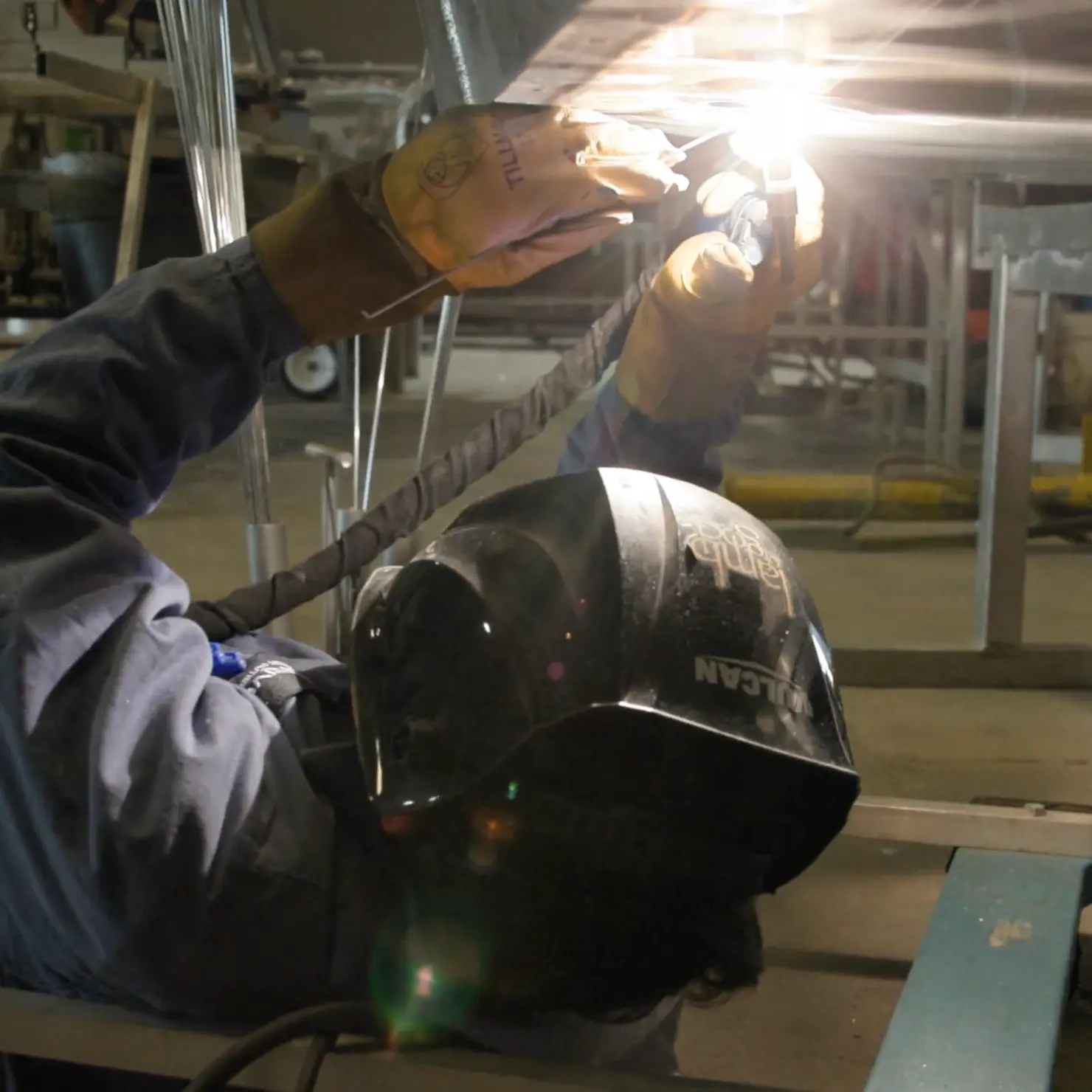 Welder wearing a protective helmet and gloves performing welding on a metal surface indoors.