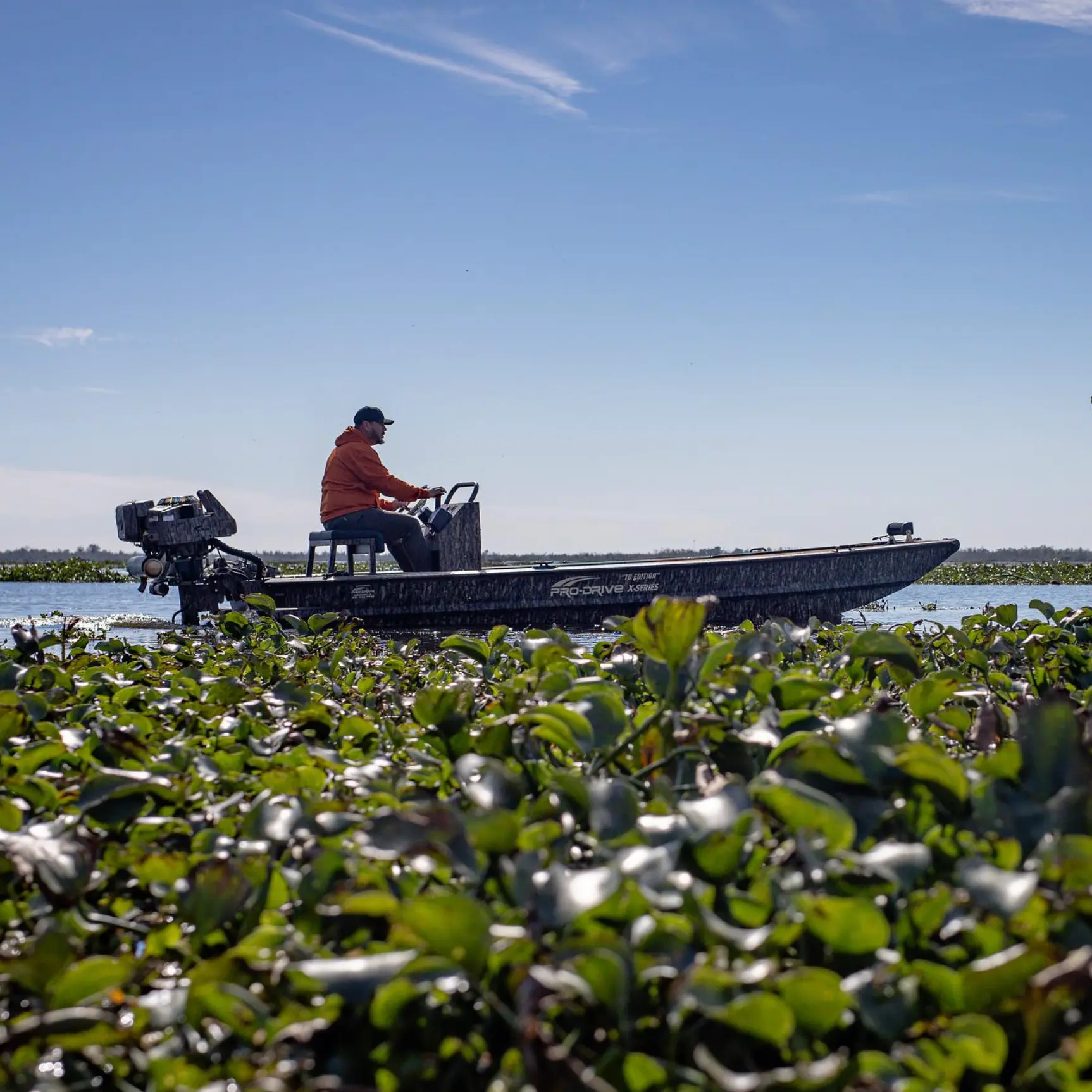 Man in an orange jacket sitting on a Pro-Drive boat navigating through water covered with green aquatic plants under a clear blue sky.