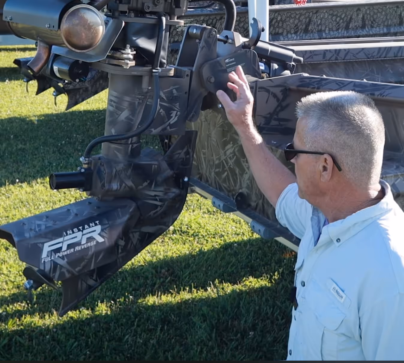 Man wearing sunglasses demonstrating the handle control of a camouflaged boat motor labeled Instant Full Power Reverse.