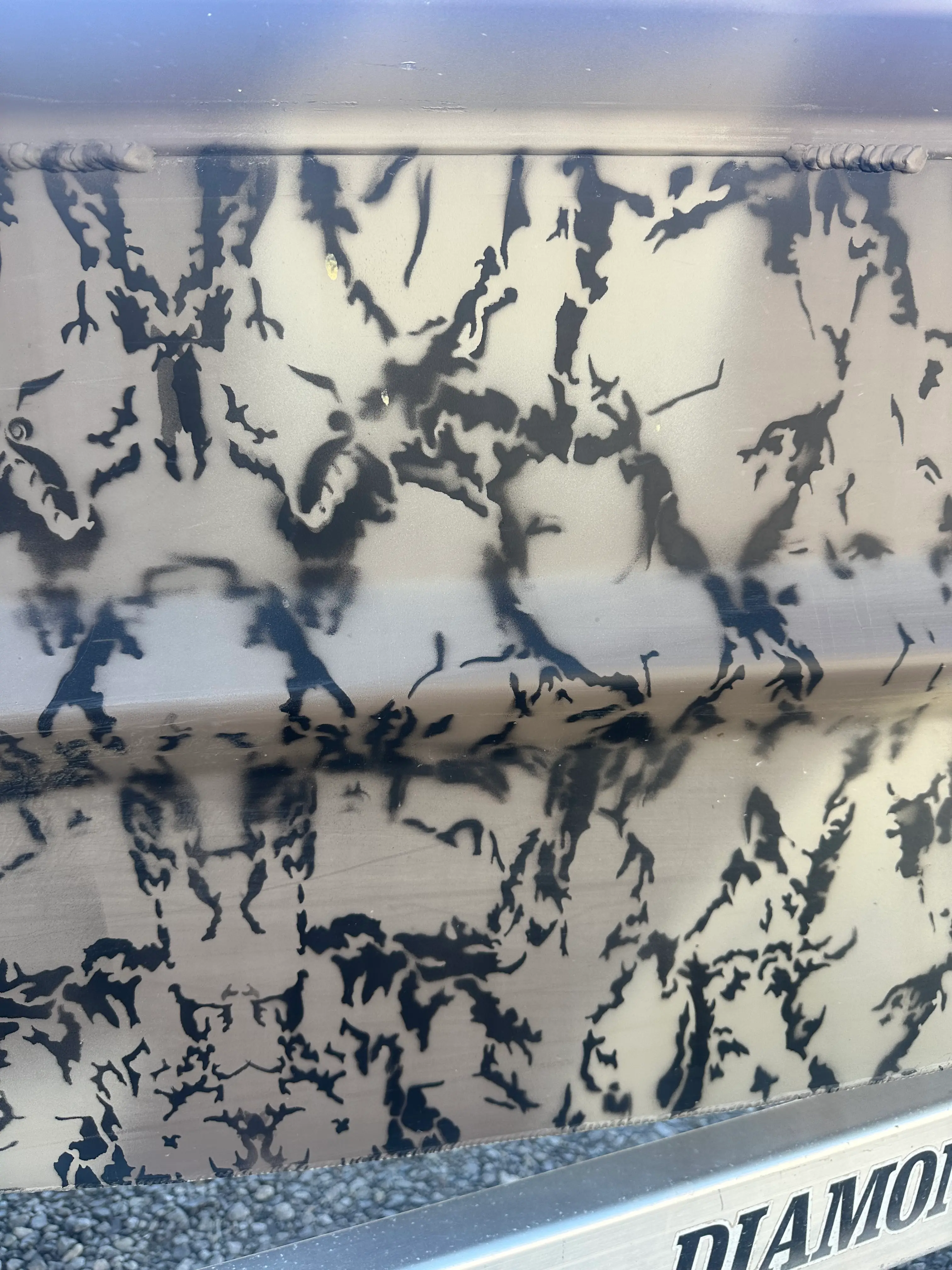 Close-up of a camouflage pattern with black abstract shapes on a beige surface, part of a metal structure with 'DIAMOND' text partially visible on a beam below.