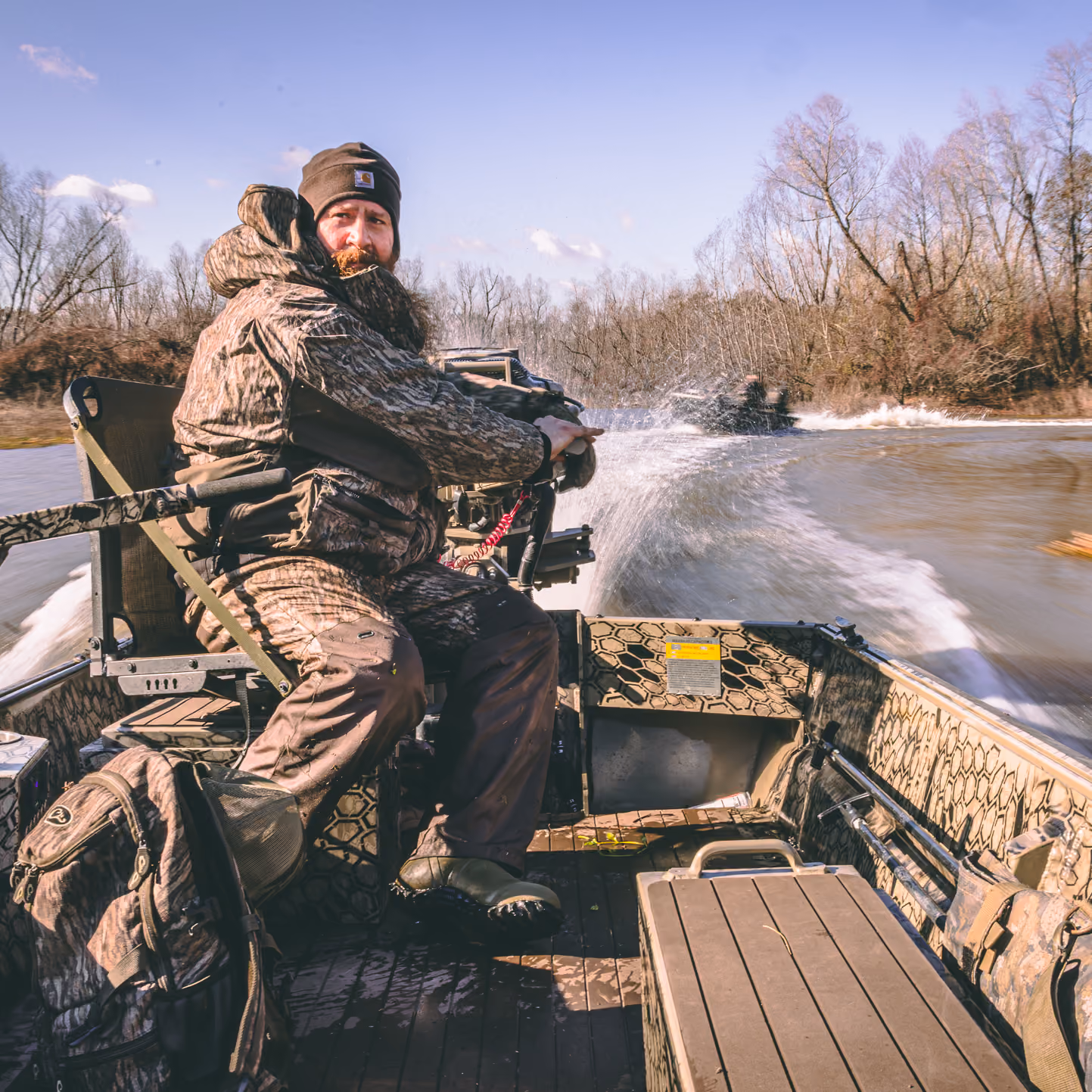 Man in camouflage gear steering a motorized boat on a river with trees in the background.