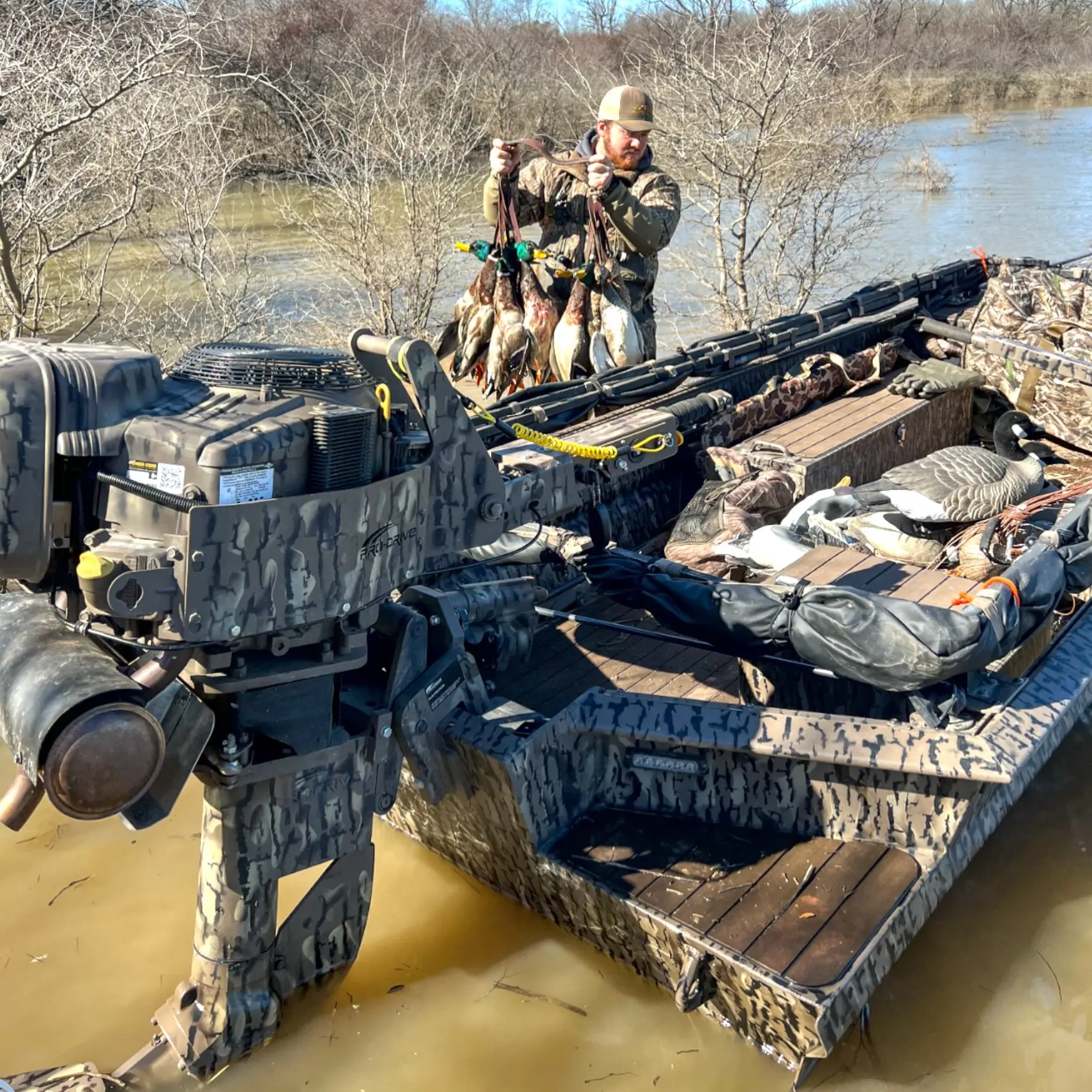 Man on a camouflage hunting boat with X40 motor holding a string of ducks in a flooded wooded area.