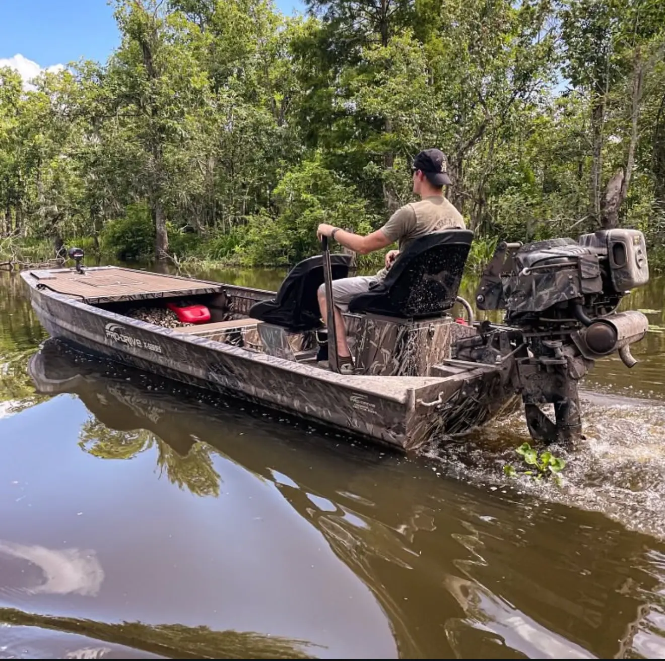 Man steering a camouflage boat with X40 motor on a calm river surrounded by trees under a partly cloudy sky.