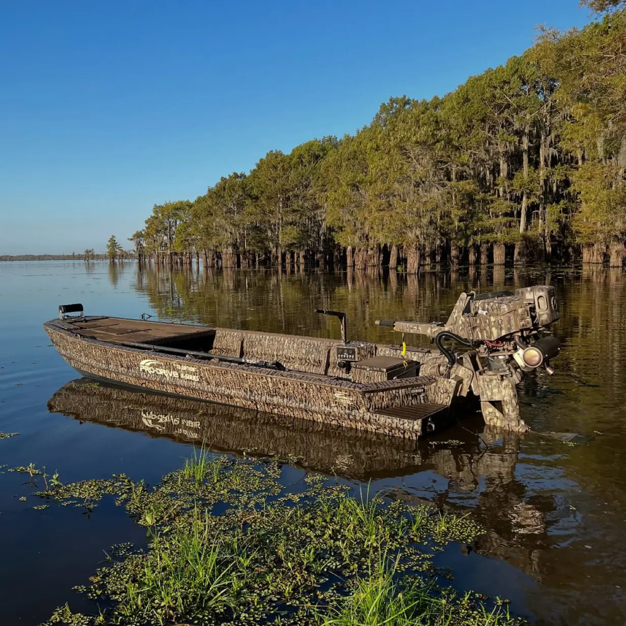 Camouflaged fishing boat with X40 motor floating on calm water near a forested shoreline under clear blue sky.