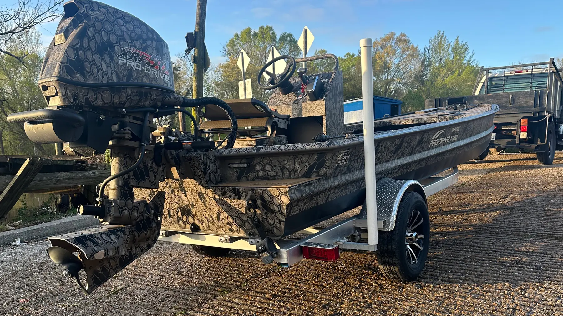 Camouflaged fishing boat on a trailer parked on a gravel path with trees and a truck in the background.