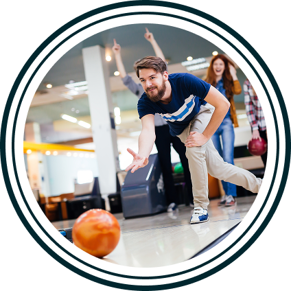 Man bowling a ball down a lane while friends cheer him on in the background at a bowling alley.