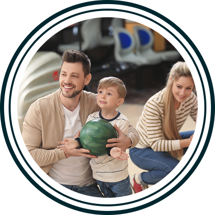 Man and young boy holding a bowling ball, with a woman smiling in the background at a bowling alley.