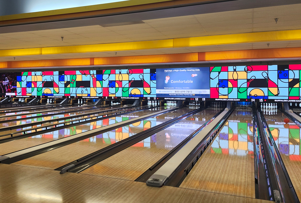 Empty bowling lanes with colorful abstract designs and a hearing aid ad on screens above the pins.