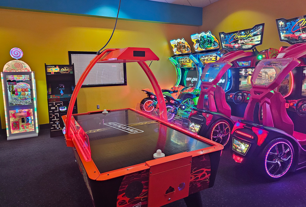 Arcade room with a red air hockey table, motorcycle racing games, and a colorful claw machine.