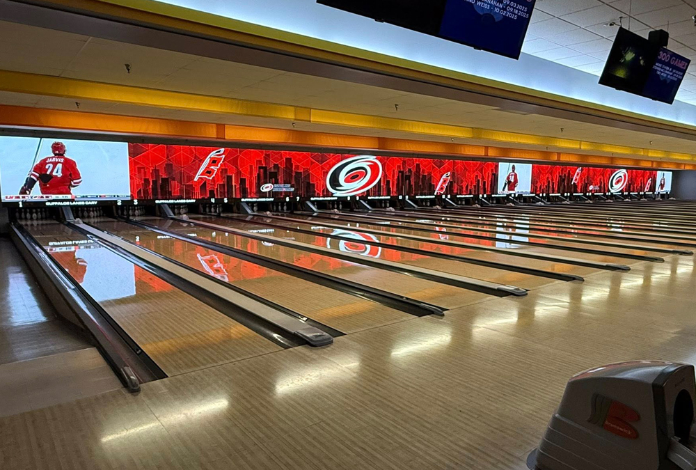 View of empty bowling lanes with red and black decor and screens showing hockey images above pins.