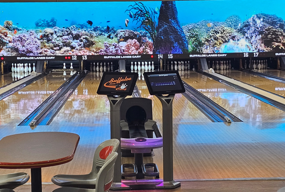 Bowling lanes with pins set, score monitors, chairs, tables, and a large ocean-themed mural above lanes.