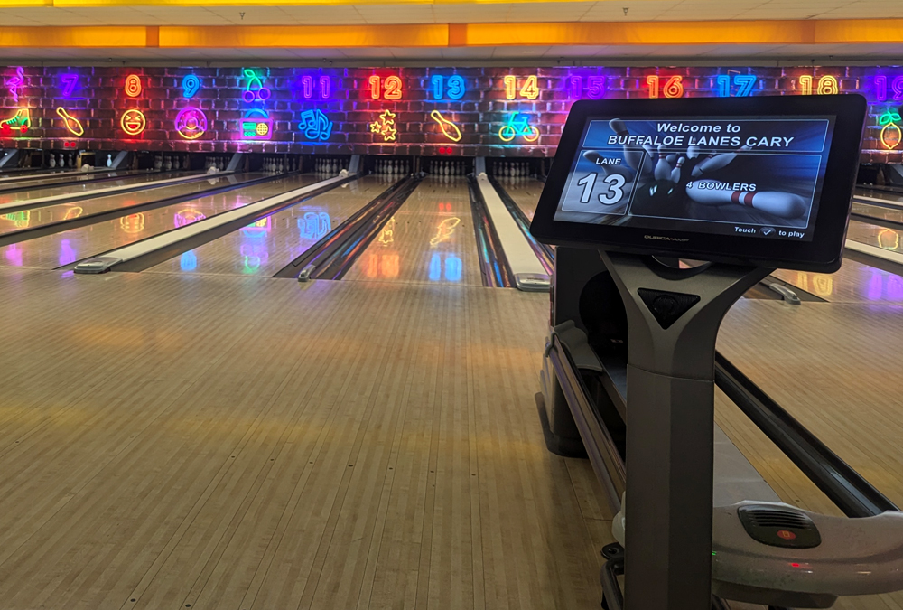 View of bowling lanes with neon signs and a digital scoring screen showing lane 13 at Buffaloe Lanes Cary.