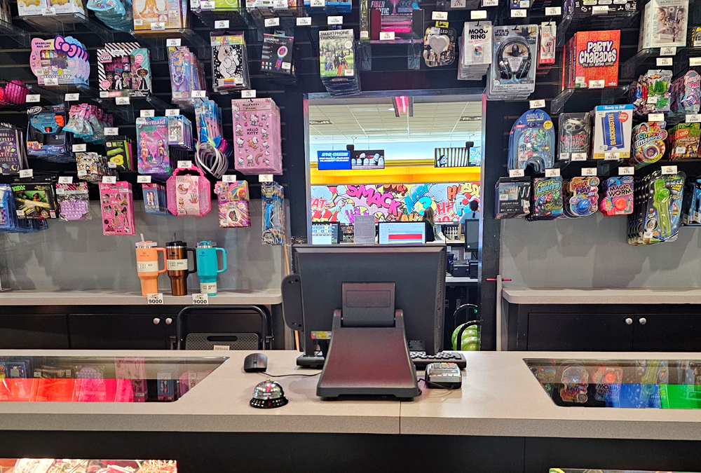 Store checkout counter with hanging toys, drink tumblers, and colorful shelves behind the register.