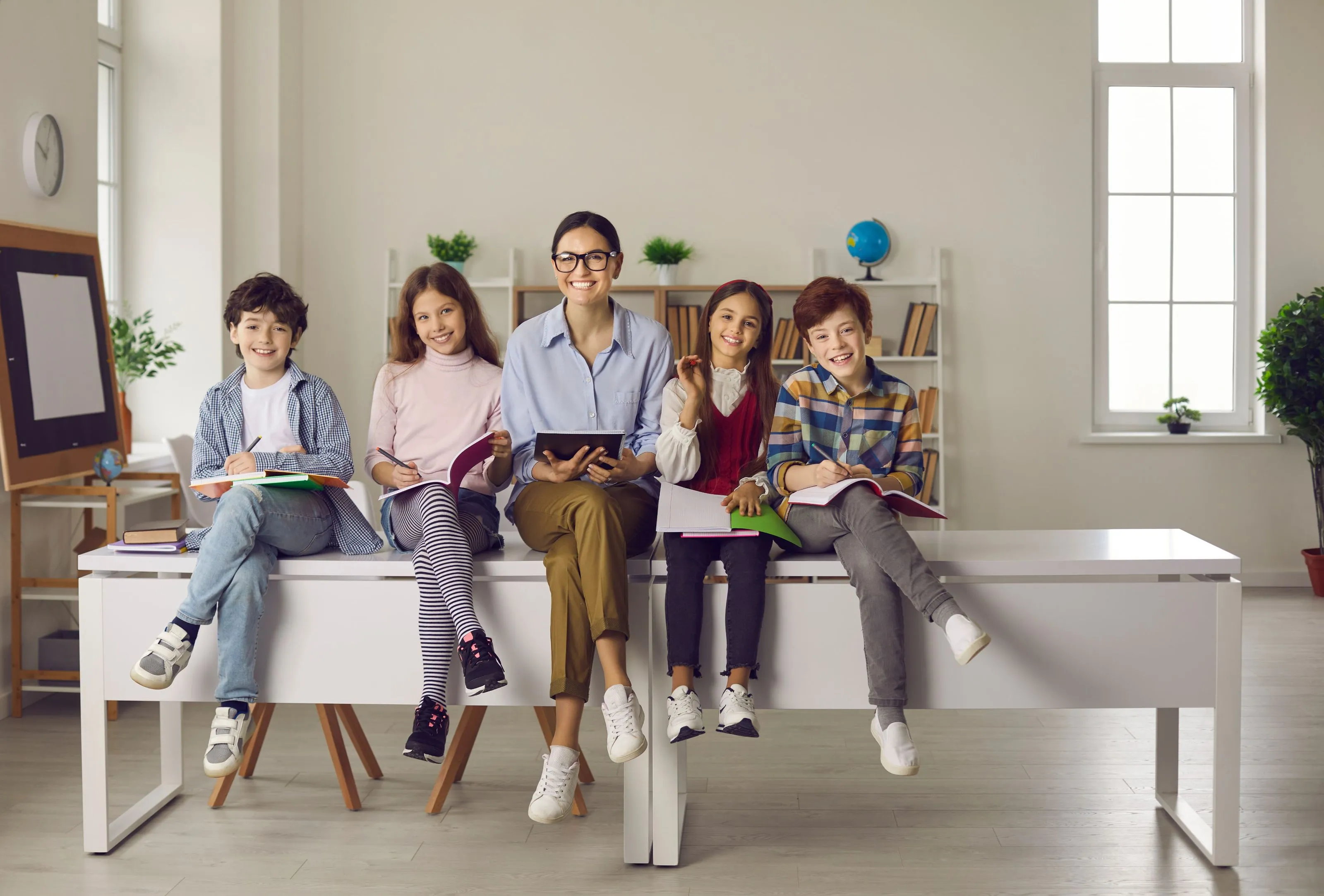 Nauczycielka siedząca z grupą uśmiechniętych dzieci w klasie, wszyscy trzymają książki.