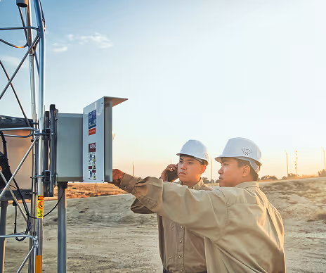 Two construction workers wearing white helmets inspecting an electrical control panel outdoors near a metal structure at sunset.