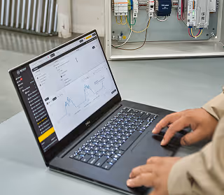Person using a laptop displaying a data monitoring dashboard with graphs and control panels, beside an open electrical control box.