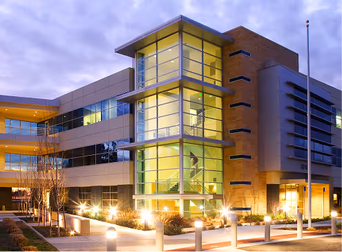 Modern three-story office building with illuminated glass staircase and exterior lights at dusk.