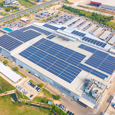 Aerial view of a large industrial building with multiple solar panels installed on its roof surrounded by parking lots and roads.