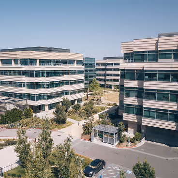 Modern office buildings with glass windows surrounding a landscaped courtyard with pathways and trees under a clear blue sky.