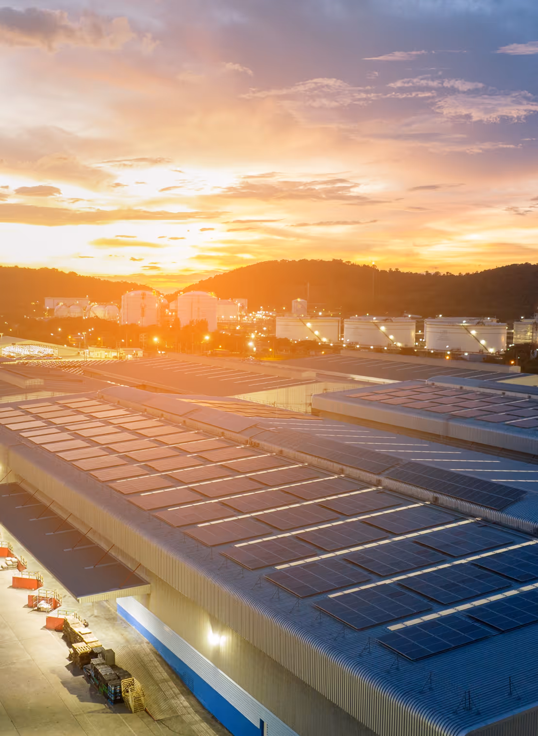 Industrial buildings with solar panels on the roofs at sunset with hills in the background and storage tanks illuminated.
