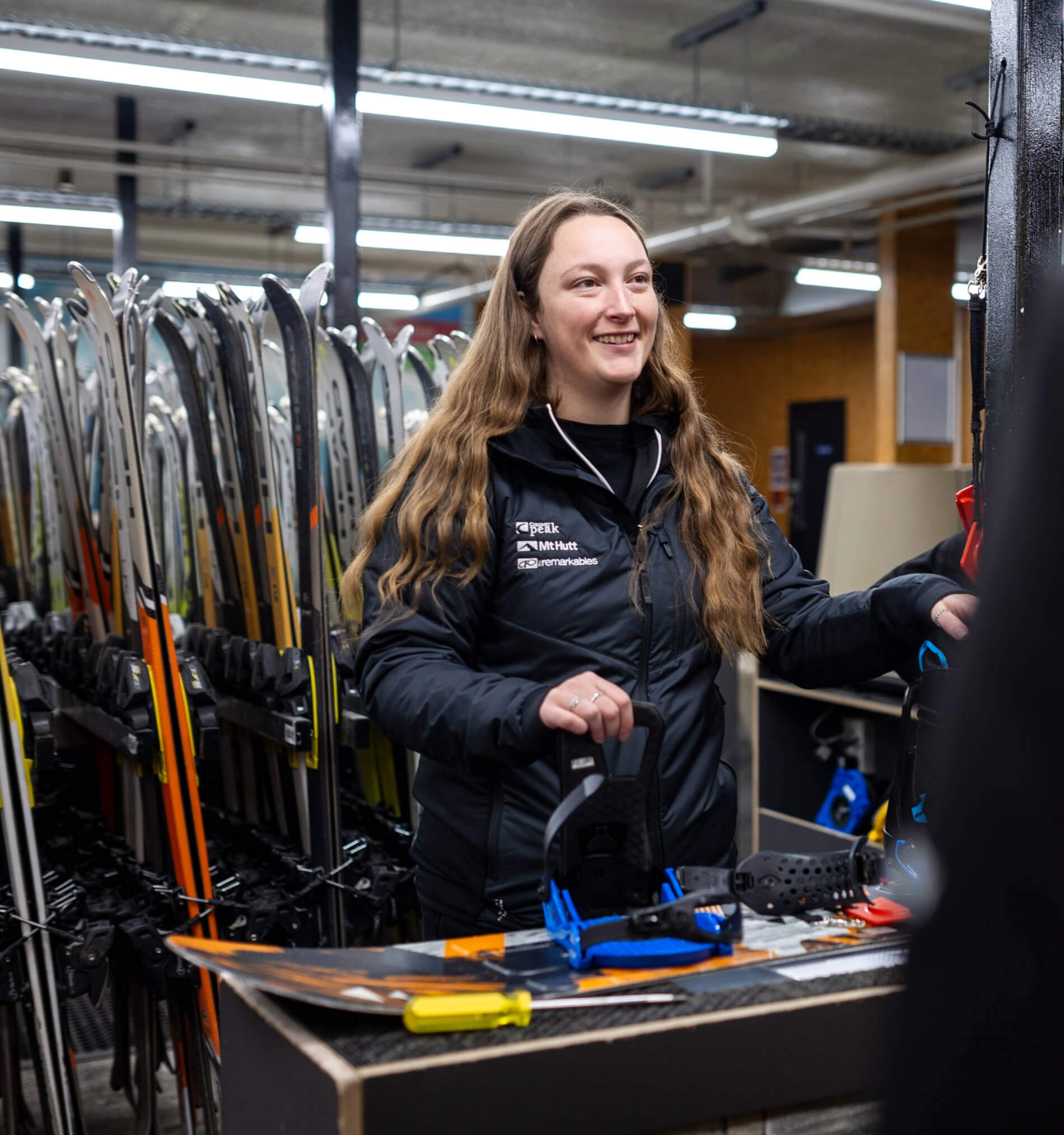 Woman smiling and working at a ski rental counter with skis and snowboards in the background.