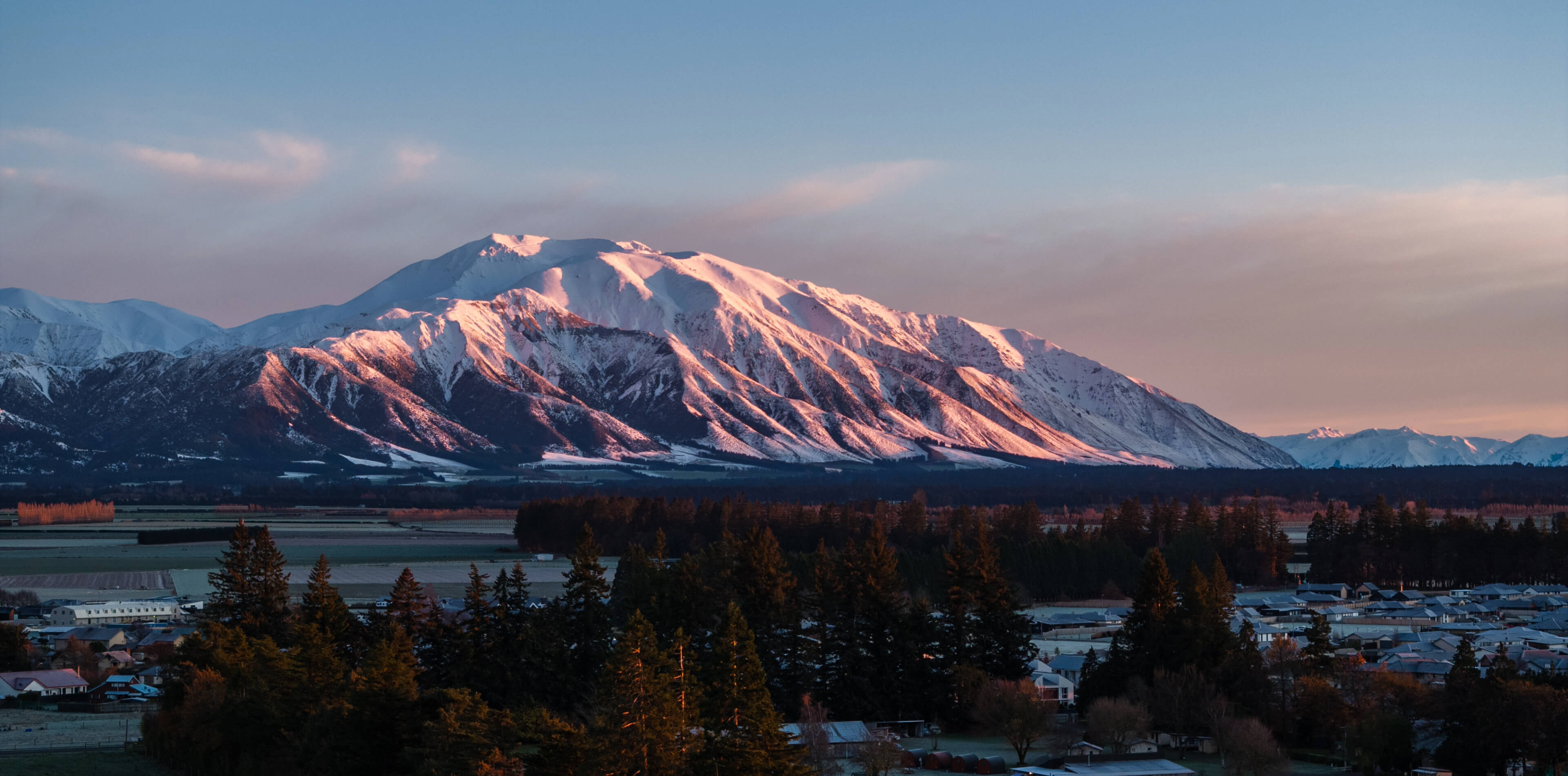 Snow-covered mountain bathed in pink sunrise light above a forest and a small town.