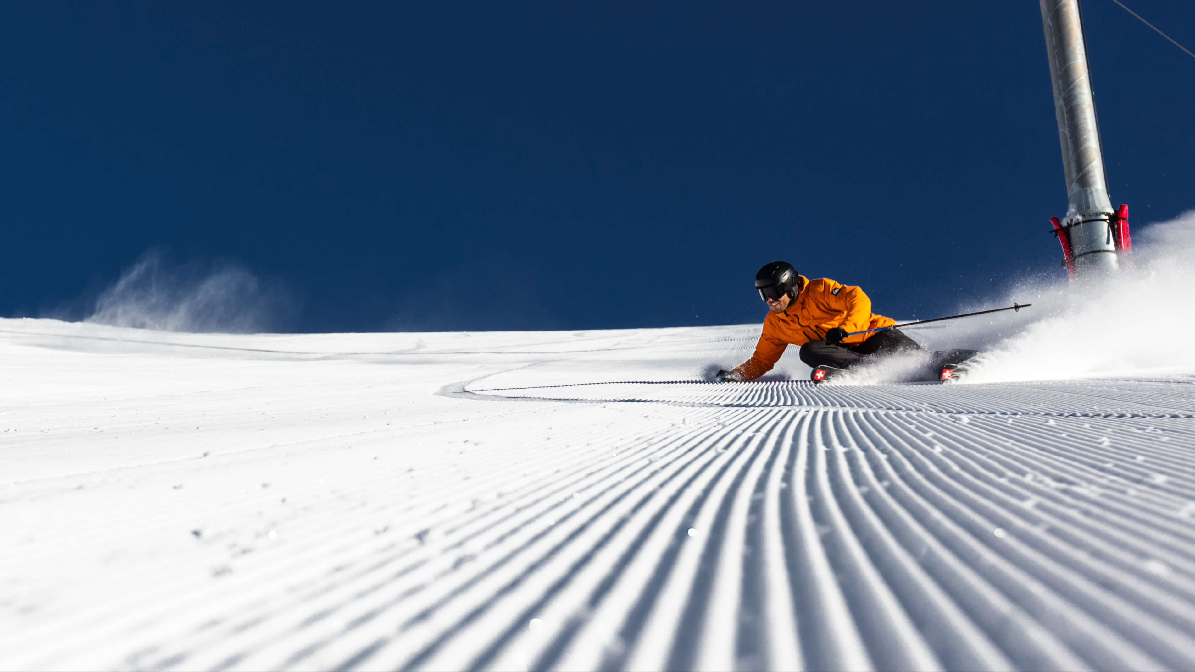 Skier in an orange jacket carving a turn on freshly groomed snow under a clear blue sky.