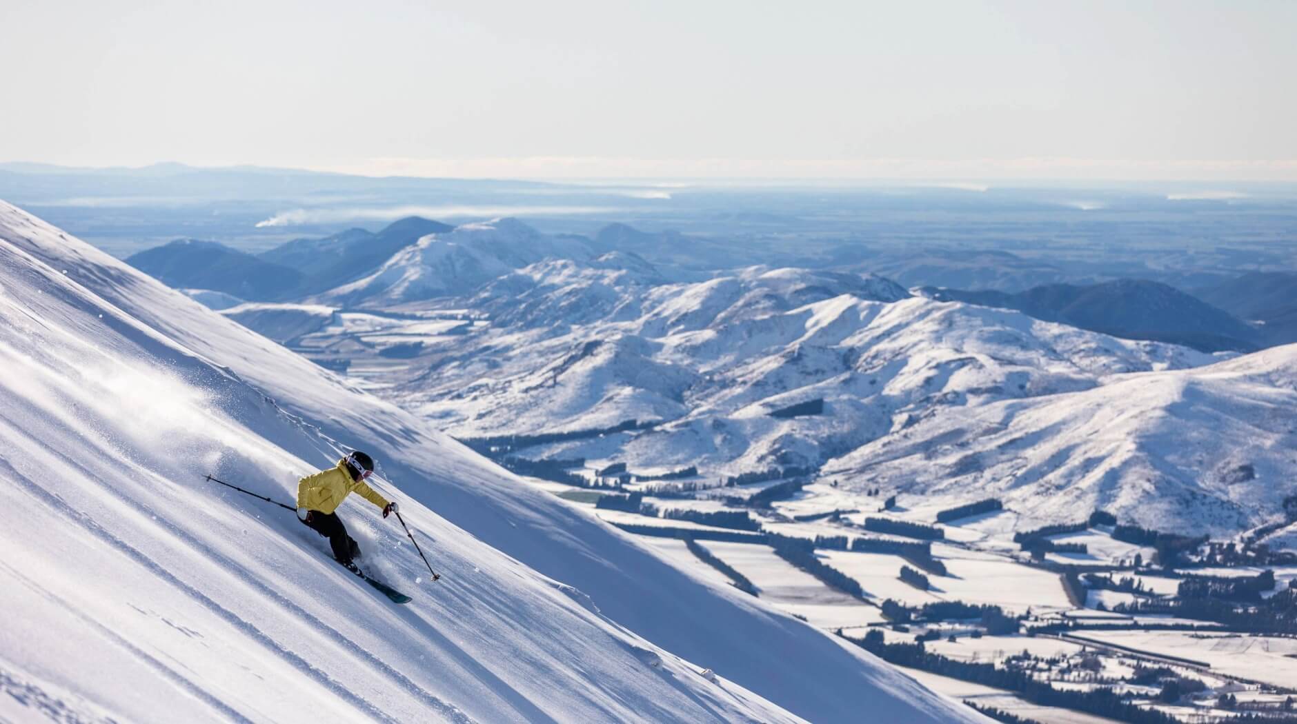Skier in a yellow jacket skiing down a snowy mountain slope with a panoramic view of snow-covered hills and valleys in the background.